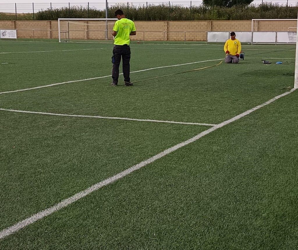 Dos trabajadores realizan labores en el césped del campo de fútbol "V Centenario" de Tagarabuena.