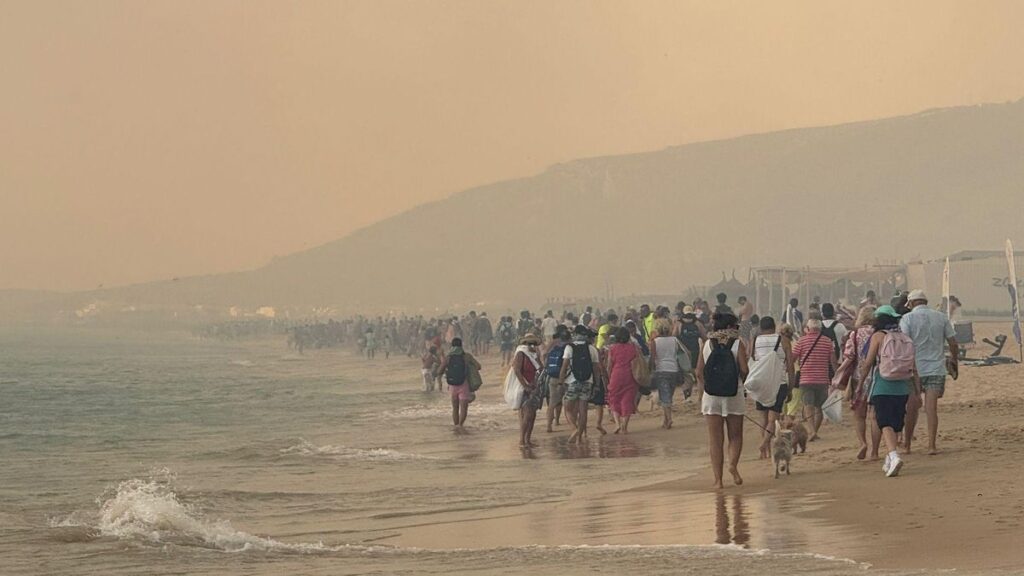 Cientos de personas caminan por la orilla de la playa dejando atrás la zona del incendio