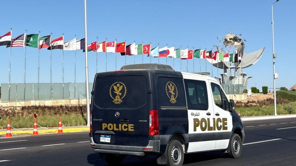 Un furgón policial pasa junto a las banderas de los países participantes en la cumbre organizada por Egipto en Sharm el Sheij para acoger la firma oficial del acuerdo de alto el fuego para Gaza.