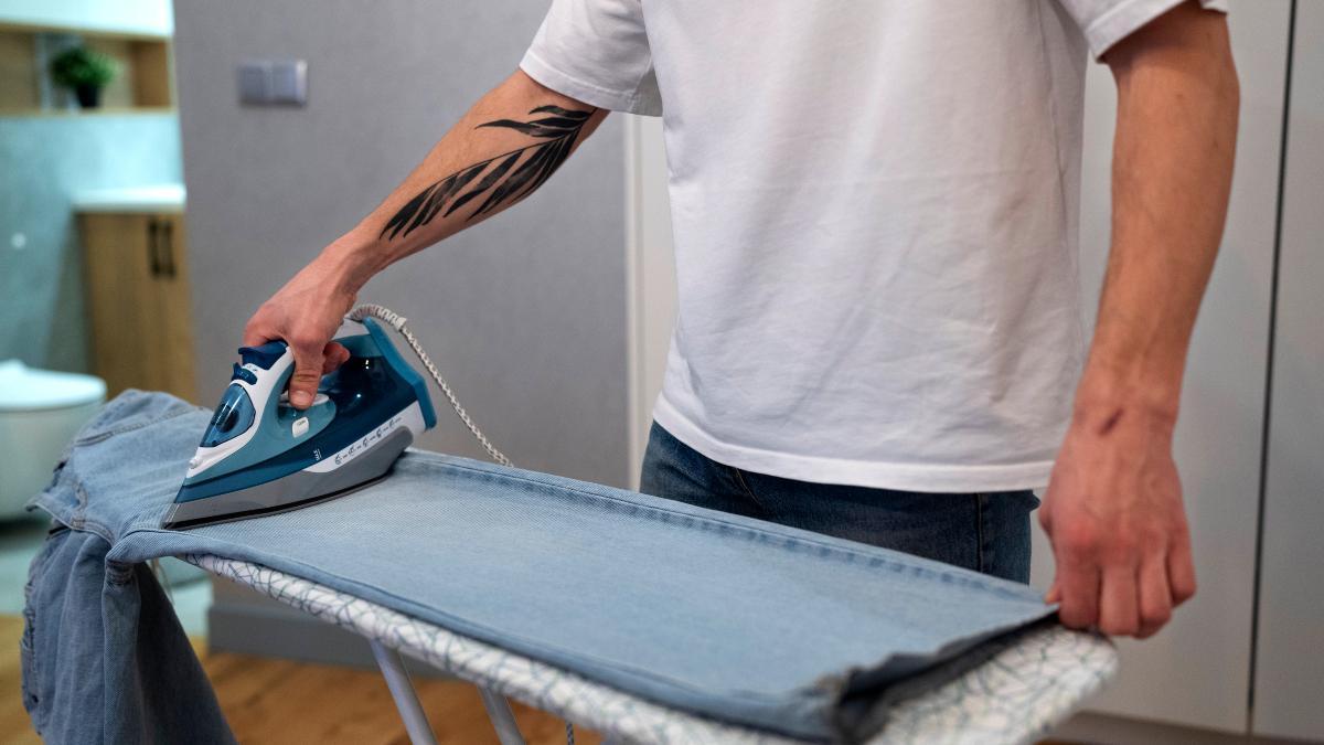 Un hombre usando la plancha para estirar unos pantalones vaqueros