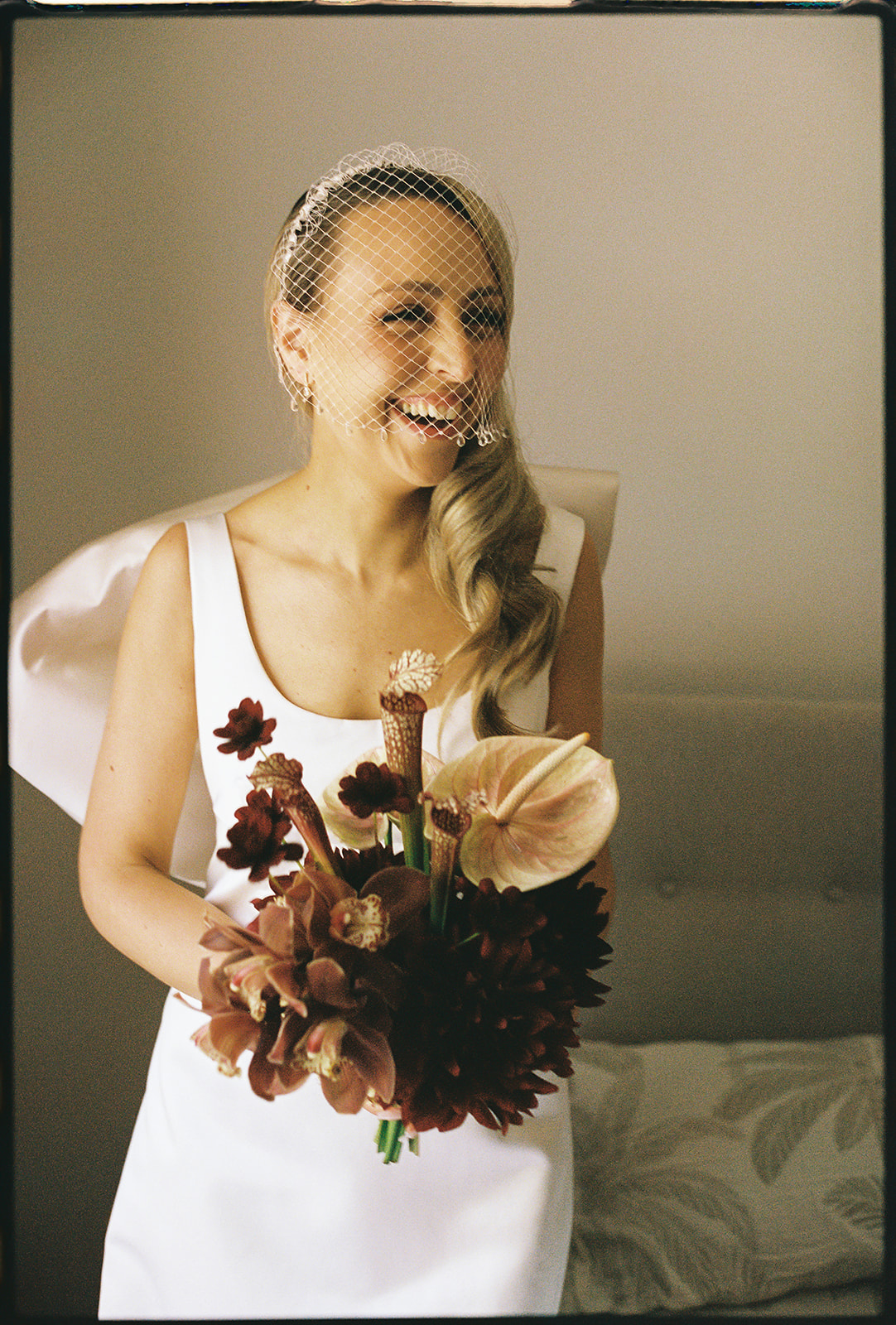 mariée enjouée portant une robe de mariée en soie personnalisée avec un nœud dans le dos et un voile en forme de cage à oiseaux, tenant un bouquet de mariée bordeaux et blush par The Beautiful Bunch – Mariage dans un vignoble de Melbourne à Marnong Estate photographié par Kinship par Kristy