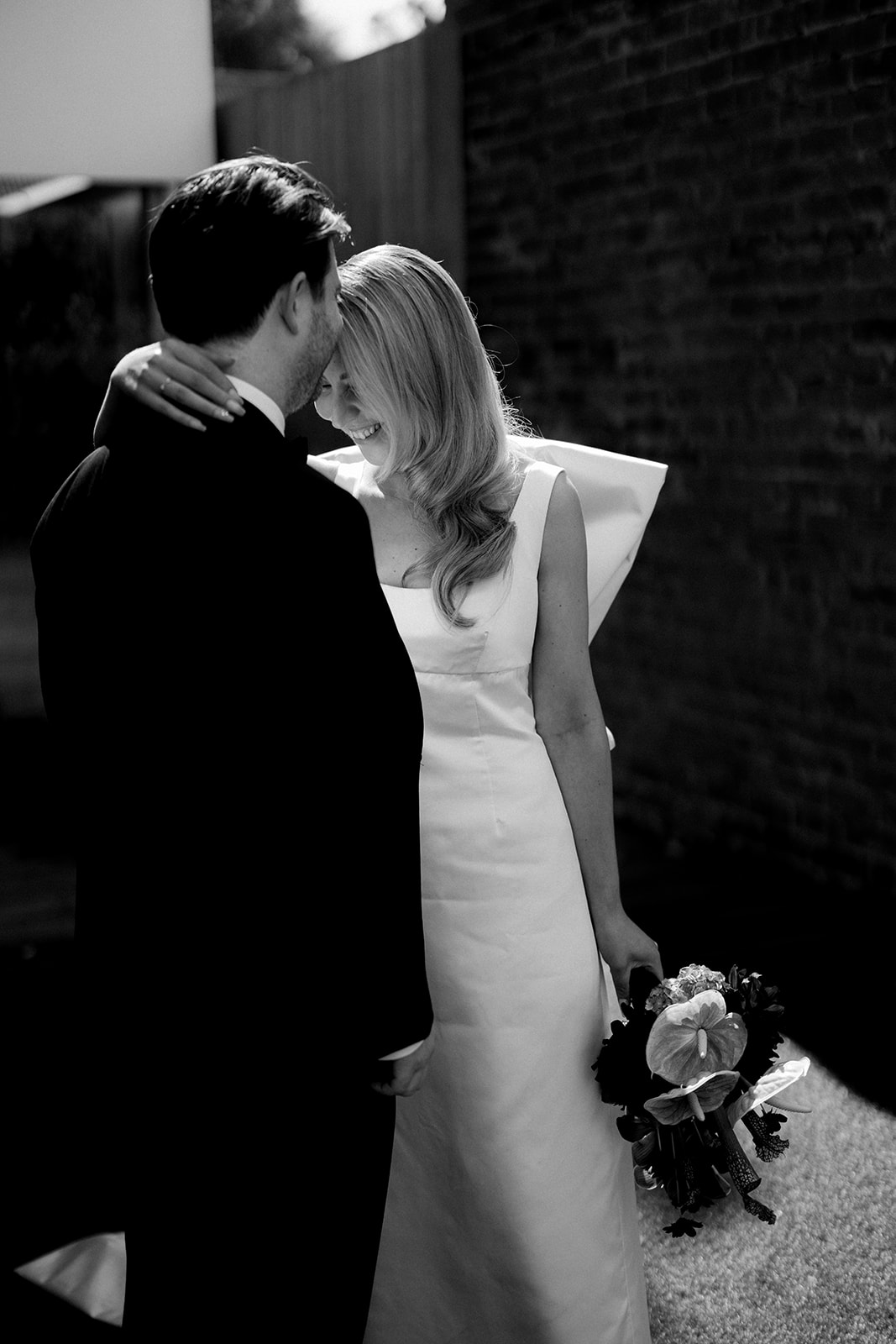 Portrait de couple romantique en noir et blanc des mariés riant ensemble dans une ruelle à Northcote, Melbourne — mariée en robe de soie conçue sur mesure tenant un bouquet de mariée bordeaux par The Beautiful Bunch, photographie de Kinship par Kristy