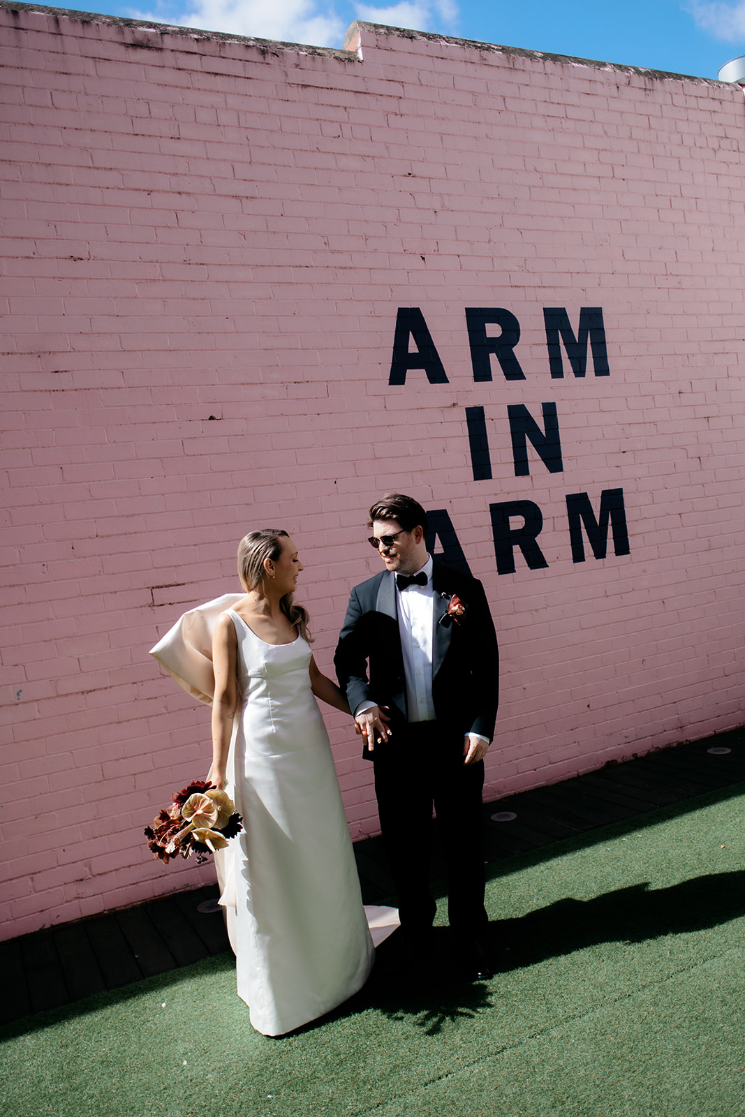 Mariée et marié se tenant la main devant un rose "Bras dessus bras dessous" fresque murale à Northcote, Melbourne – mariée portant une robe de mariée en soie personnalisée avec nœud au dos, marié en smoking noir – photographie de mariage par Kinship by Kristy